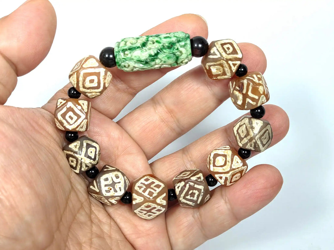 Ancient Tibetan dzi bracelet with geometric-etched honey-brown agate beads, black onyx spacers, and a carved green jadeite focal bead, held in an open palm