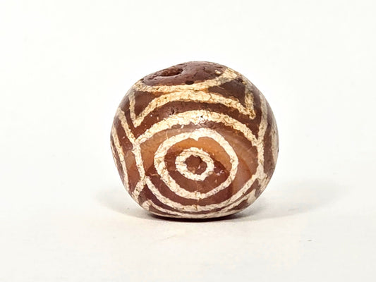 Ancient carnelian four eye heavens gate dzi bead, warm orange-brown agate with pale cream etched eye and stripe motifs, round form showing natural surface calcification and age patina, unretouched photo by Ancient Dzi Shop