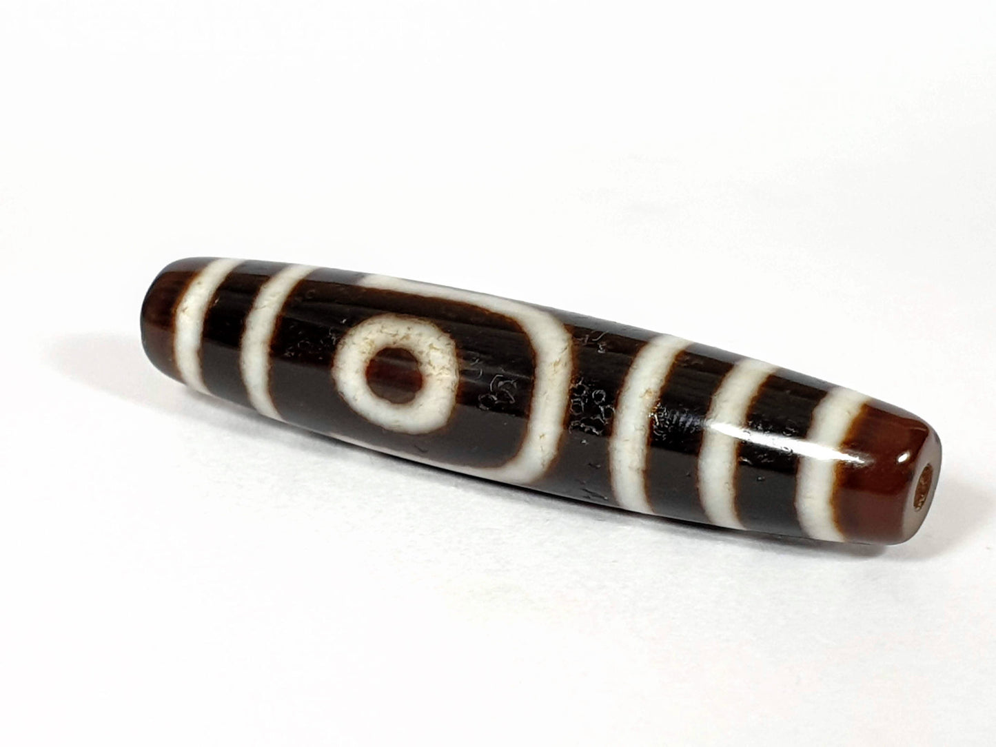 V-070226-2E2 dzi bead, diagonal view showing drill hole, brown ground, unretouched photo.
