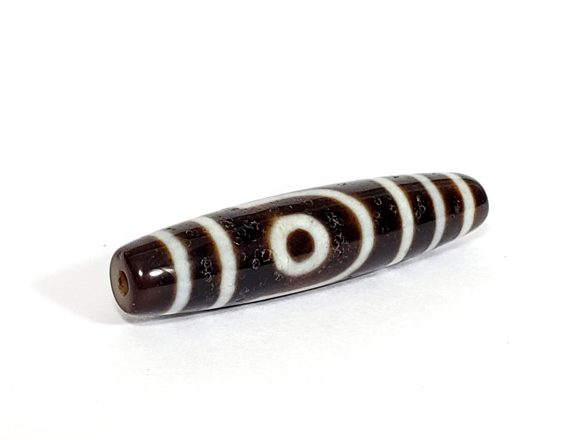 V-070226-2E dzi bead, diagonal view showing drill hole, dark brown ground, unretouched photo.
