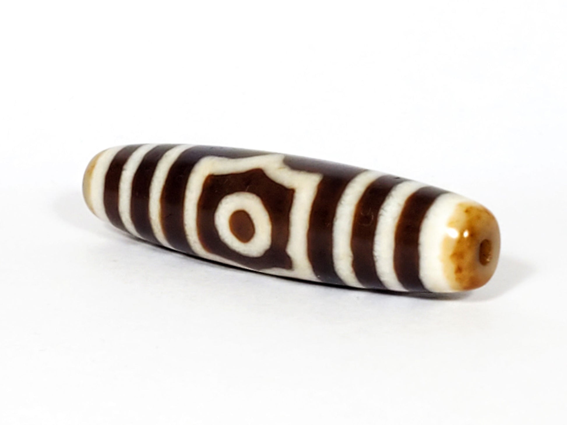 Three-quarter view of antique three eye dzi At-053126-3E, warm honey-tan end cap with drill hole, octagonal-frame eye visible, chocolate-brown torpedo body, unretouched