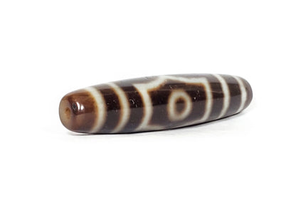 V-070226-ThE-003 dzi bead, diagonal view showing drill hole, dark brown ground, unretouched photo.