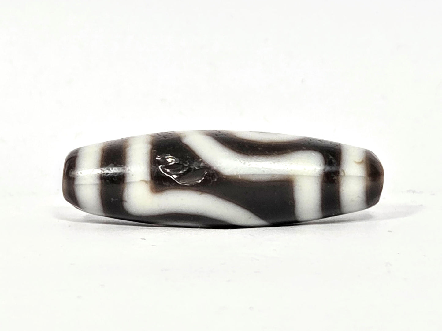 Side profile of antique two eye dzi bead At-010726-2E3, torpedo body, near-black agate, cream vertical stripes, medicine digs visible, 38.6mm × 12.5mm