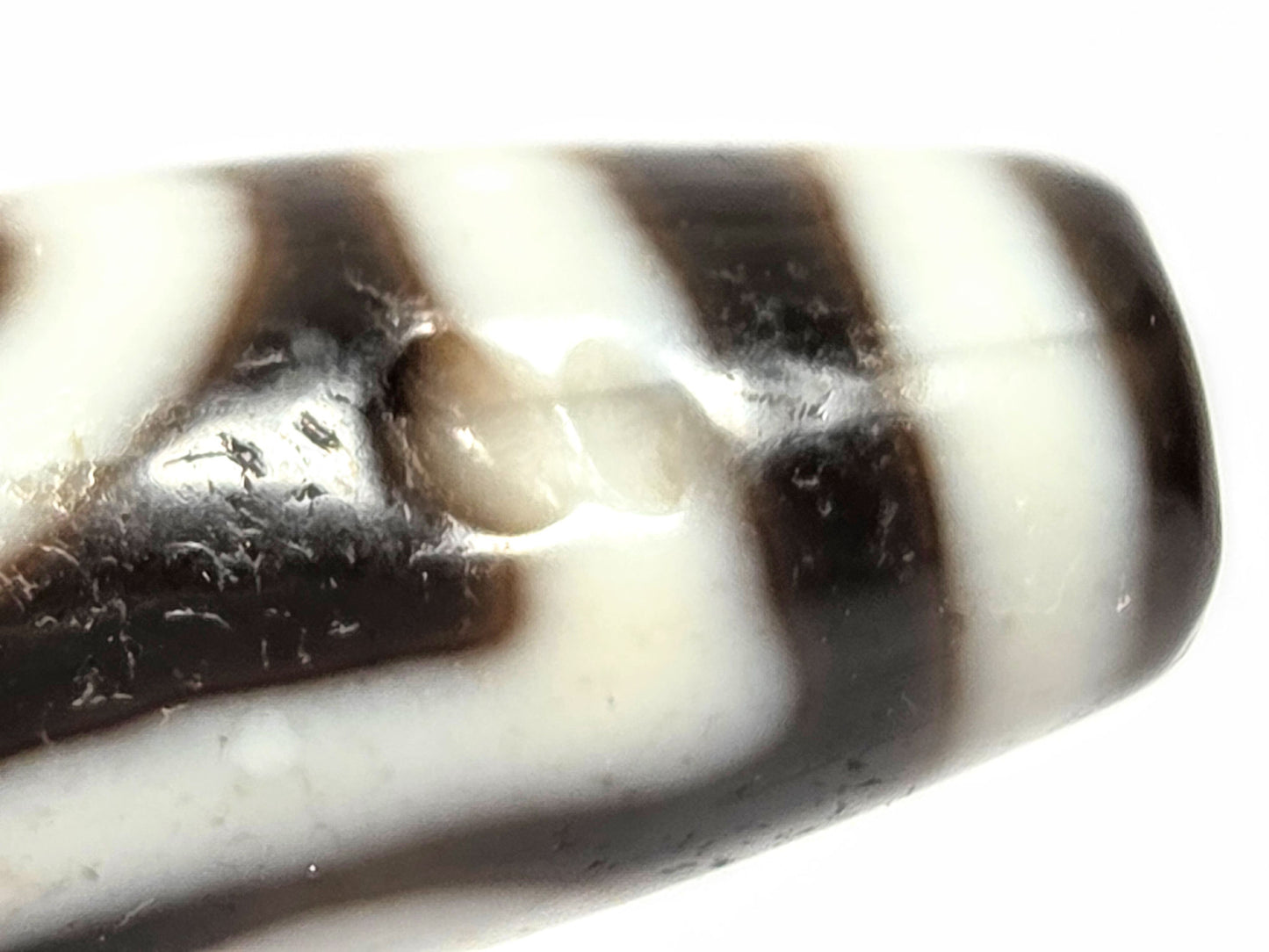Close detail of antique two eye dzi bead At-010726-2E3 showing medicine dig on near-black agate surface, authentic age marking, unretouched photography