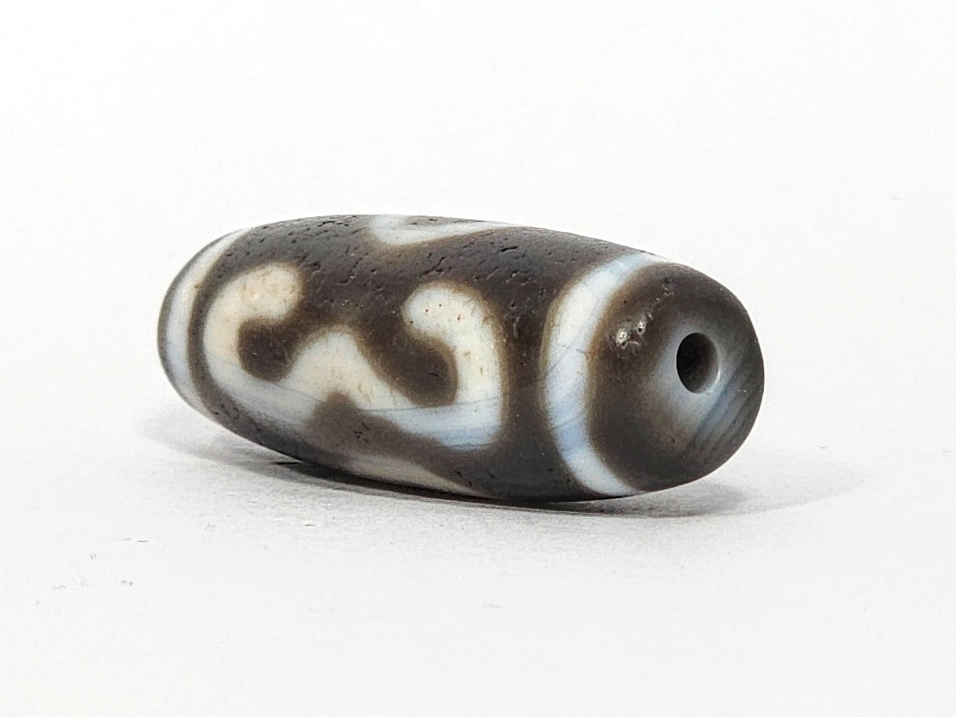 Three-quarter view of antique Double Bat Two Eye dzi At-060725-DB2E2, grey-brown end cap with drill hole, bat wing S-curves visible on body, matte aged agate surface, unretouched