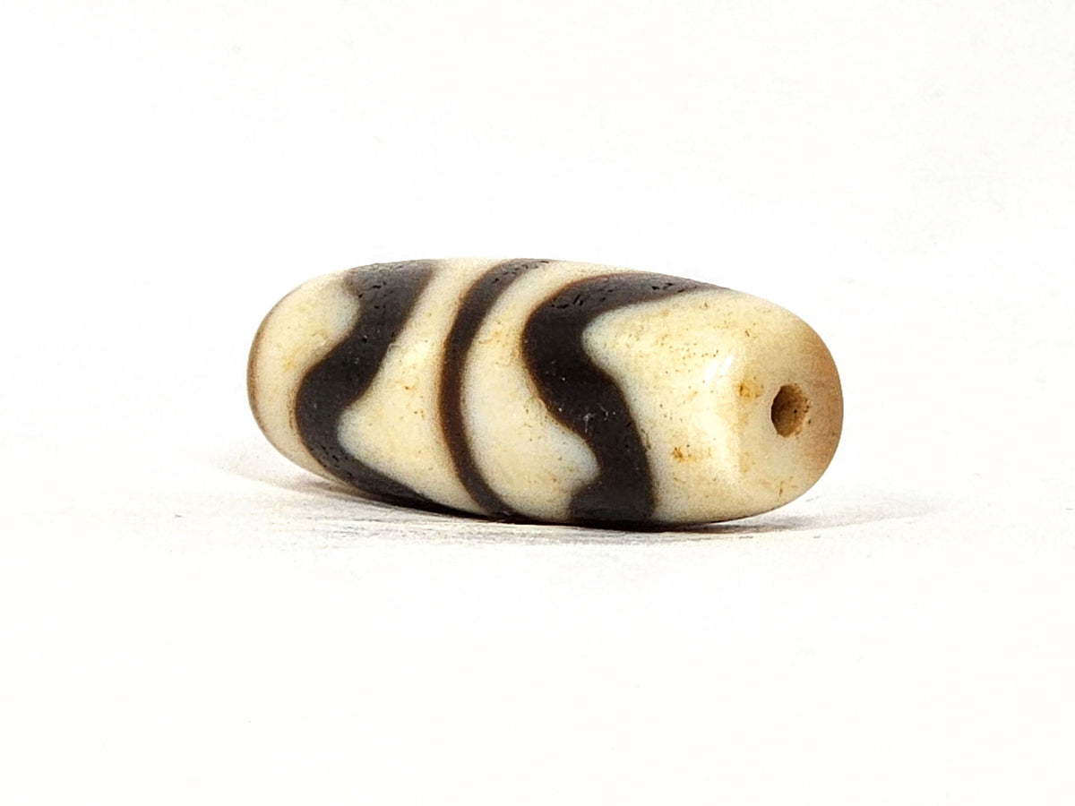 Three-quarter view of antique Double Wave dzi At-072325-DW5, warm brown end cap with drill hole, wave motif visible on compact torpedo body, 28.4mm × 10.6mm, unretouched
