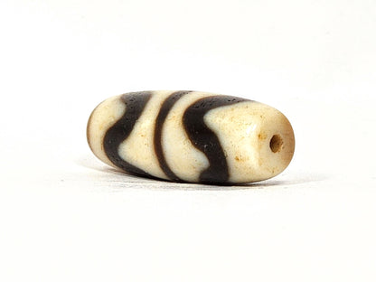 Three-quarter view of antique Double Wave dzi At-072325-DW5, warm brown end cap with drill hole, wave motif visible on compact torpedo body, 28.4mm × 10.6mm, unretouched