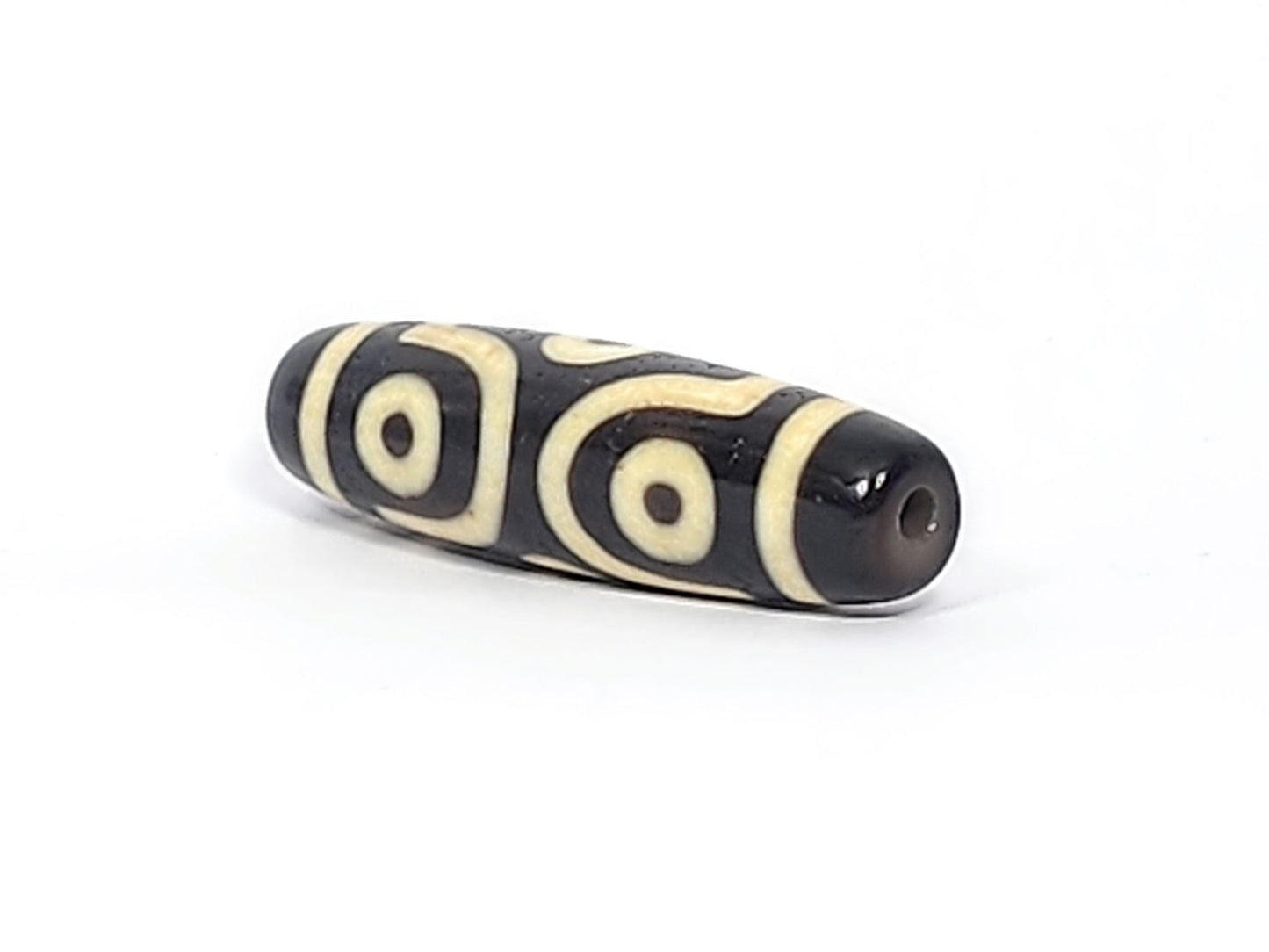 Three-quarter view of antique six eye dzi At-042925-6E2, pitch-black end cap with drill hole, two C-ring eyes and scroll visible, elongated torpedo body, unretouched