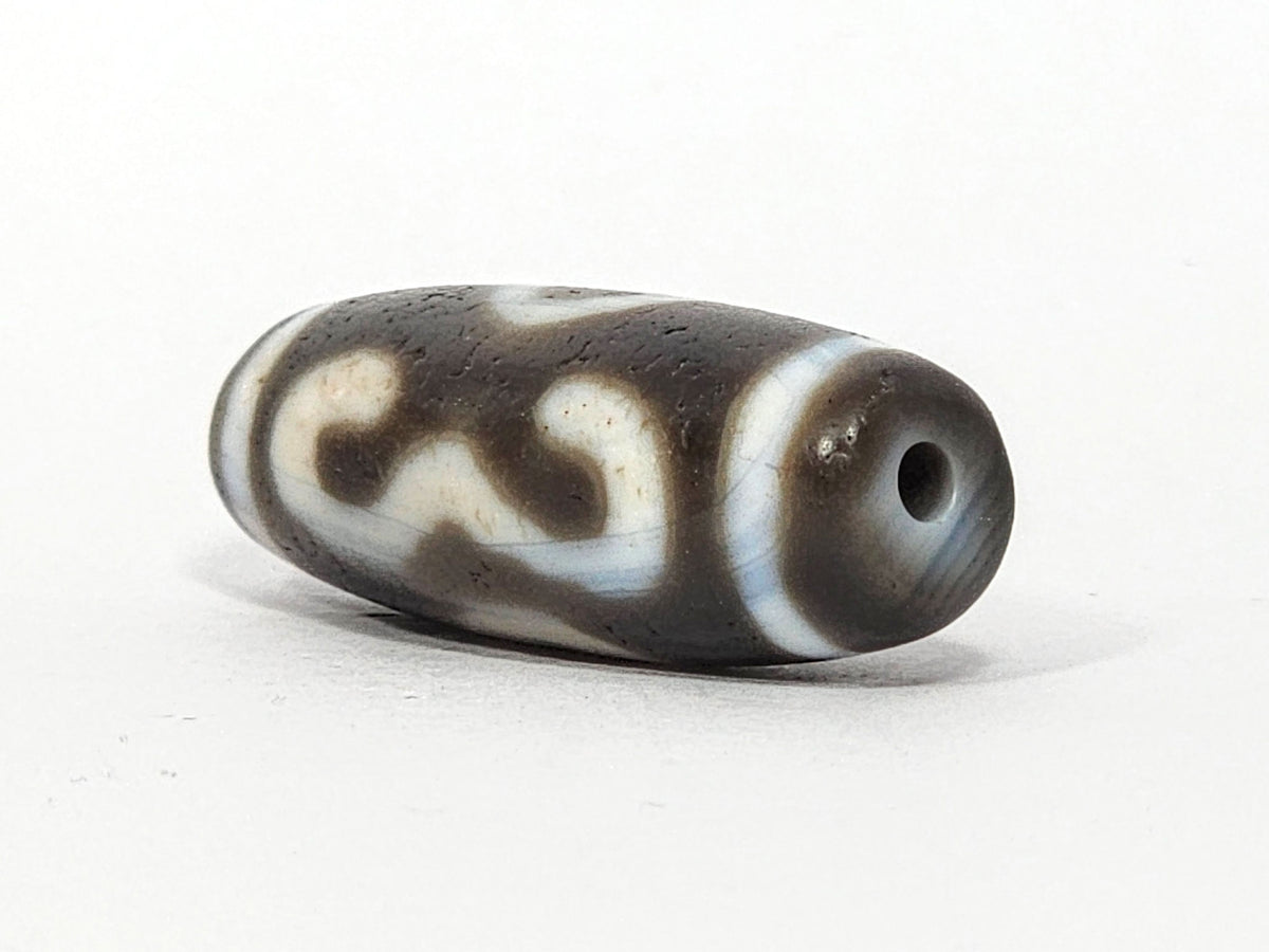 Opposite end of antique Double Bat Two Eye dzi At-060725-DB2E2, grey-brown end cap, drill hole, bat S-curve motif wrapping rectangular barrel, aged agate surface texture, unretouched