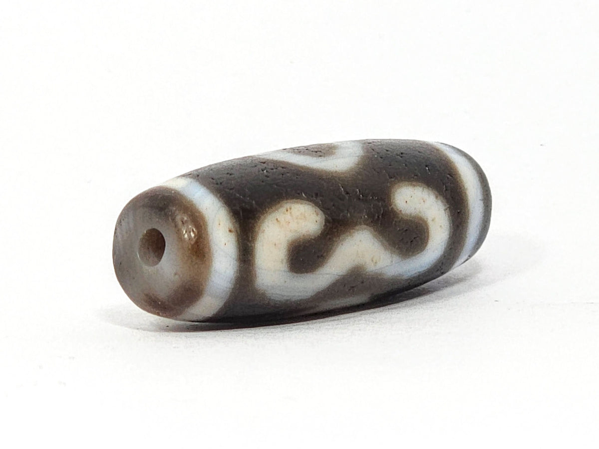 Three-quarter view of antique Double Bat Two Eye dzi At-060725-DB2E2, grey-brown end cap with drill hole, bat wing S-curves visible on body, matte aged agate surface, unretouched
