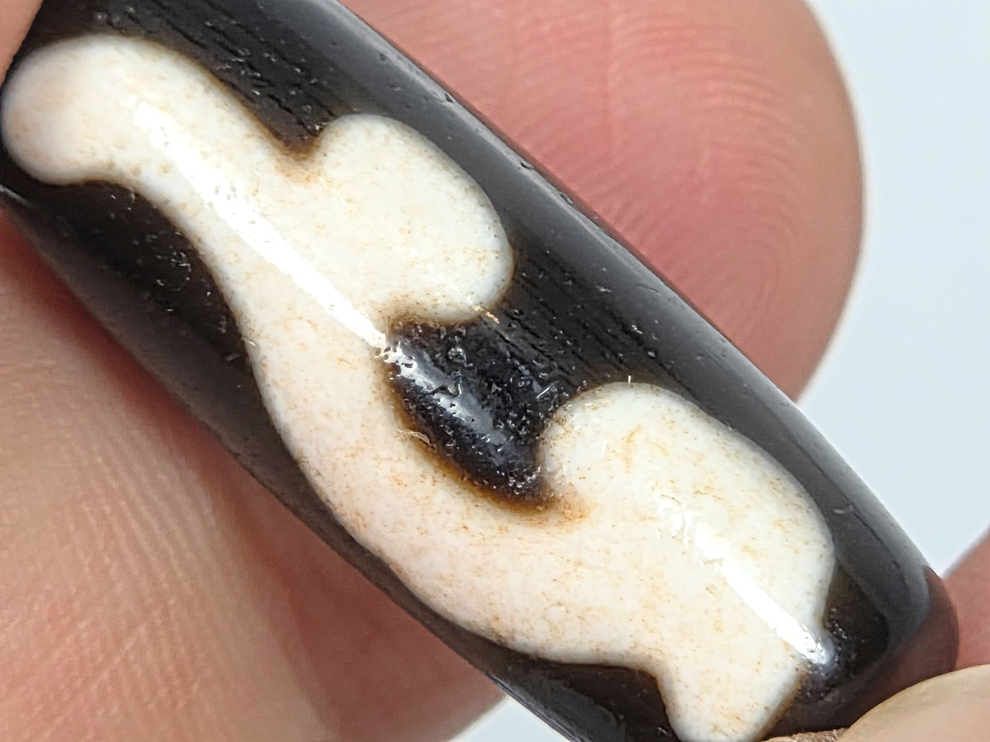 Reverse face of a 1980s Ruyi dzi bead with deep black inclusion and cream-white patterning, held between two fingers to show scale and surface texture