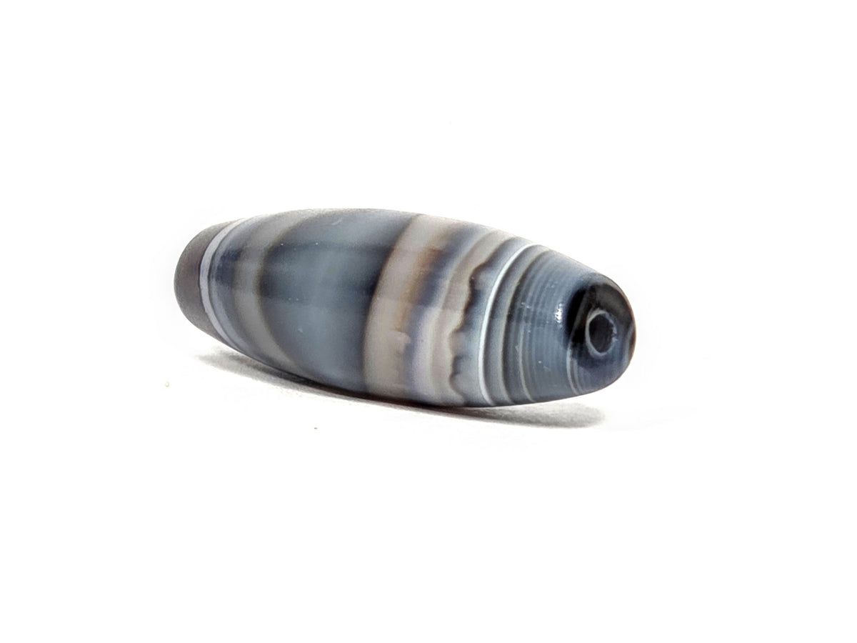 N-N-080926-1E6 natural one eye dzi bead 50.5×15.8mm, angled drill-end view showing dark end cap, cream shoulder stripe, and blue-grey body, unretouched