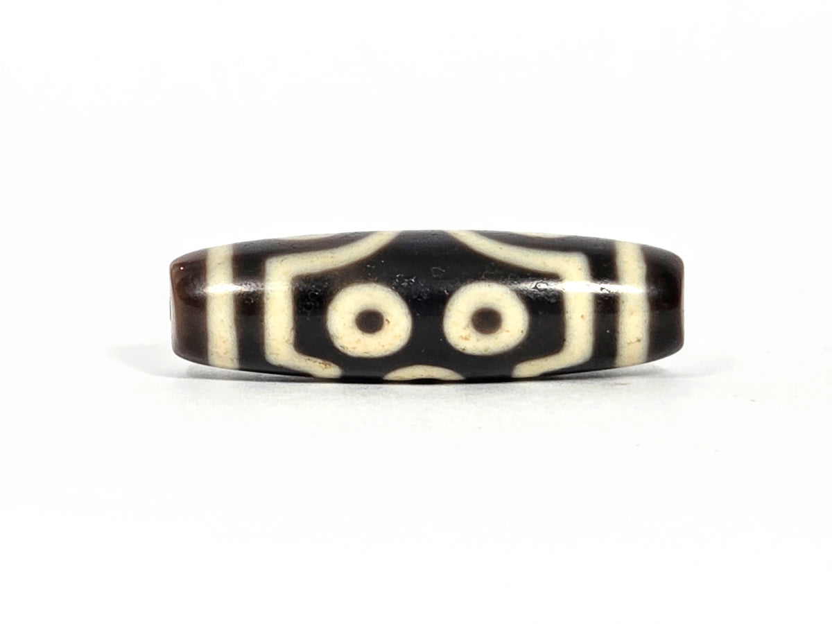 Fourth lateral profile of antique seven-eye dzi bead (AT-092826-7E) showing two eyes in paired arrangement with angular bracket framing elements, dark brown-black and cream-white agate